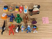 Overview of all 19 assorted character toys laid out on a wood surface showing variety including Kung Fu Panda, several Smurfs, Sonic, a green robot, penguin, and other figures.