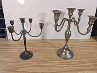 Two metal candelabras displayed on a wooden surface, showing brass candelabra with green patina and stainless steel candelabra.