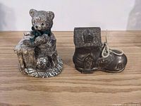 View of two metal piggy banks on a wooden surface: a teddy bear with a green ribbon sitting on a base holding a book and soccer ball, and a shoe-shaped bank with a house structure on top.