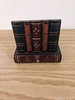 Front view of wooden pencil holder resembling four books vertically stacked on a closed book base, with painted spine and cover details in green, brown and gold.