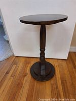 Front view of the antique wooden plant stand showing round top, turned pedestal column, and round base on hardwood floor background.
