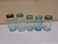Five vintage mason jars arranged side by side on white fabric background showing size variations and metal lids.