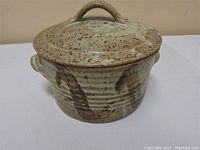 Handmade ceramic casserole dish with lid on top, showing the speckled beige and brown glaze with dark brown accents.