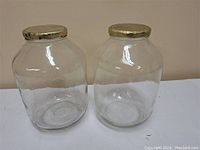 Two large clear glass pantry jars side by side on a table against a beige wall, showing full front view.