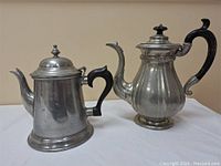 Front view of two pewter tea or coffee pots on white cloth background