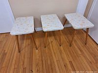 Three matching retro end tables lined up side by side on wooden floor, showing floral tops and wooden legs.