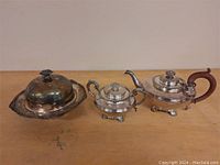 Set of three silver plated items: covered butter dish with tray, teapot, and sugar bowl all on wooden surface against plain background.