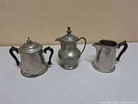 Three vintage pewter items placed side by side on a beige background, showing front and side profiles.