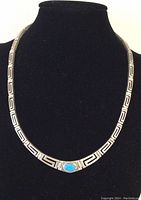 Full necklace displayed on black bust to show length and overall design with Greek key links and central turquoise stone.