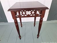 Front angled view of vintage wooden plant stand showing square tabletop, decorative lattice sides, turned legs, and minor surface marks.
