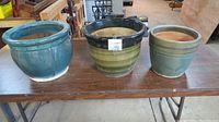 Set of three ceramic garden pots: blue, green/black, and green/blue, placed on a wooden table in a workshop setting.