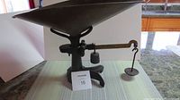 Full side view of antique weight scale showing cast iron base, steel weight pan, and brass weighing beam with hanging counterweight, placed on a counter.