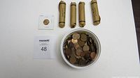 Photo of a white bowl holding a large quantity of Canadian pennies, four brown paper rolls of pennies, and one single penny in a cardboard coin holder placed on a flat surface with a MaxSold lot number card.