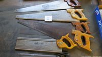 Photo showing six carpenter saws laid out on a surface, highlighting handles and blades’ length and condition with one saw heavily rusted.