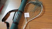 Fishing rod holder case and fishing net lying on a wooden surface, showing the entire length of the case and the net.