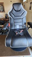 Front view of black and gray XRocker gaming chair with armrests and ergonomic design, cables and remote placed on seat.