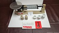 Photo showing Ohaus triple beam balance scale with metal pan, five calibration weights, and instruction booklet on white background.