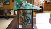 Wide shot of Roughriders pennant leaned on top of framed mirror with vintage quarterback illustrations, on a table in a basement setting.
