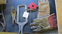 Photo showing two pairs of welding gloves, one large locking C-clamp, red triangular magnet holder, hammer with ribbed handle, and smaller locking plier clamp