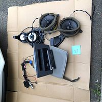 Multiple car stereo components including the Kenwood face-off head unit, wiring harness, two round car speakers, and mounting brackets displayed on a cardboard surface.