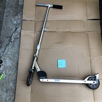 Razor kick scooter upright on cardboard surface showing front wheel, handlebar with foam grip on one side, metal frame and deck