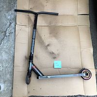 Madgear black kick scooter laid flat showing side view with visible wheel, deck and handlebar view. Foam handle grip missing on one side.