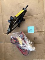 Black scissor jack placed on cardboard with bundled booster cables in plastic wrapping next to it, both tagged as Lot 038.