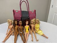 Photo showing six Barbie dolls sitting in front of a woven pink and black tote bag with silver button details
