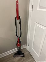 Full view of red Dirt Devil upright vacuum cleaner standing against a wall near a white door, showing power cord wrapped around the handle and clear dust compartment.