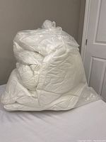 Folded white feather bed topper in clear plastic bag placed on a bed.