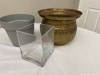 Three flower pots shown together: a grey plastic pot, a clear glass square container, and a brass pot.