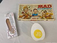 Photo shows three items: a boxed MAD Magazine game with illustrated cartoon characters on cover, a magnetic dartboard game in a white box with wooden handle photo, and a white egg-shaped kitchen timer with yellow center reading 'EGG HEAD'.