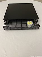 Photo of black metal mesh K-Cup coffee pod organizer drawer closed, showing solid top surface and drawer front with wire frame and mesh base, placed on a white surface.