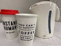 Photo showing an electric kettle alongside two coffee travel mugs, one with a red lid and 'INSTANT HUMAN JUST ADD COFFEE' text, the other with coffee-related quote.