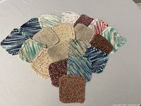 Top down view of all sixteen colorful homemade face cloths arranged on a flat surface, variety of yarn colors, primarily blues, greens, purples, browns and creams.