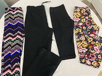 Four women's leggings laid flat on a white surface: one full length multicolor zigzag patterned pair, one full length solid black pair, one capri length solid black pair, one capri length floral patterned pair.
