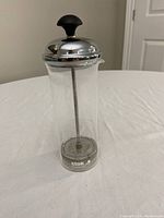 Photo of the Stok milk foamer showing clear glass container with stainless steel lid and plunger, and black knob handle on top.