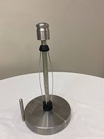 Single stainless steel paper towel holder with round weighted base and vertical rod holding tension arms and an upper handle.