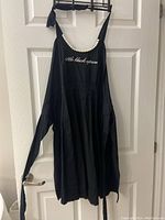 Front view of black apron hung on door showing pearl embellishments on neckline and embroidered text 'little black apron'.
