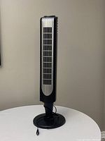 Full view of the black tall oscillating tower fan standing upright on a round base against a plain background.