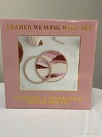 Box of framed weaving wall art kit showing the packaging and image of completed woven art in pink and brown hues.