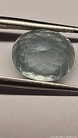 Close-up of oval cut bluish aquamarine gemstone held by tweezers showing detailed facets and color.