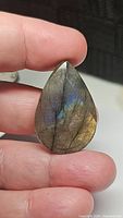 Photo of a pear-shaped labradorite gemstone showing natural iridescent flash with blue and yellow tones, held between fingers for scale.