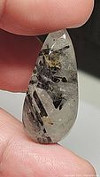 Close-up of 28.38ct pear-shaped natural black rutilated quartz cabochon showing clear quartz with needle-like black tourmaline inclusions inside and some gold flecks, held between thumb and finger