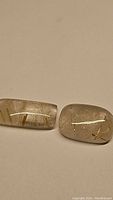 Two natural golden rutilated quartz stones with visible golden needle inclusions, lying flat on a beige surface. One stone is slightly wider and the other more elongated.