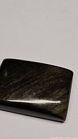 Close-up photo of natural golden sheen obsidian cabochon showing golden flash reflections on dark polished surface.