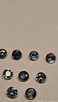 Nine round cut natural sapphires laid out on a plain background. Stones show aqua blue to teal multicolored hues, each approximately 2.5mm in diameter.