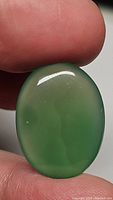 Close-up photo showing a smooth oval green onyx cabochon held between fingers, highlighting its translucency and polished surface.