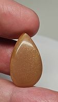 Close-up photos of a pear-shaped cabochon cut peach moonstone gemstone showing its size relative to fingers and its sparkling shimmer surface.