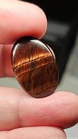 Close-up of natural red tiger eye oval cabochon held between fingers showing its polished surface and rich reddish-brown color with chatoyant bands.